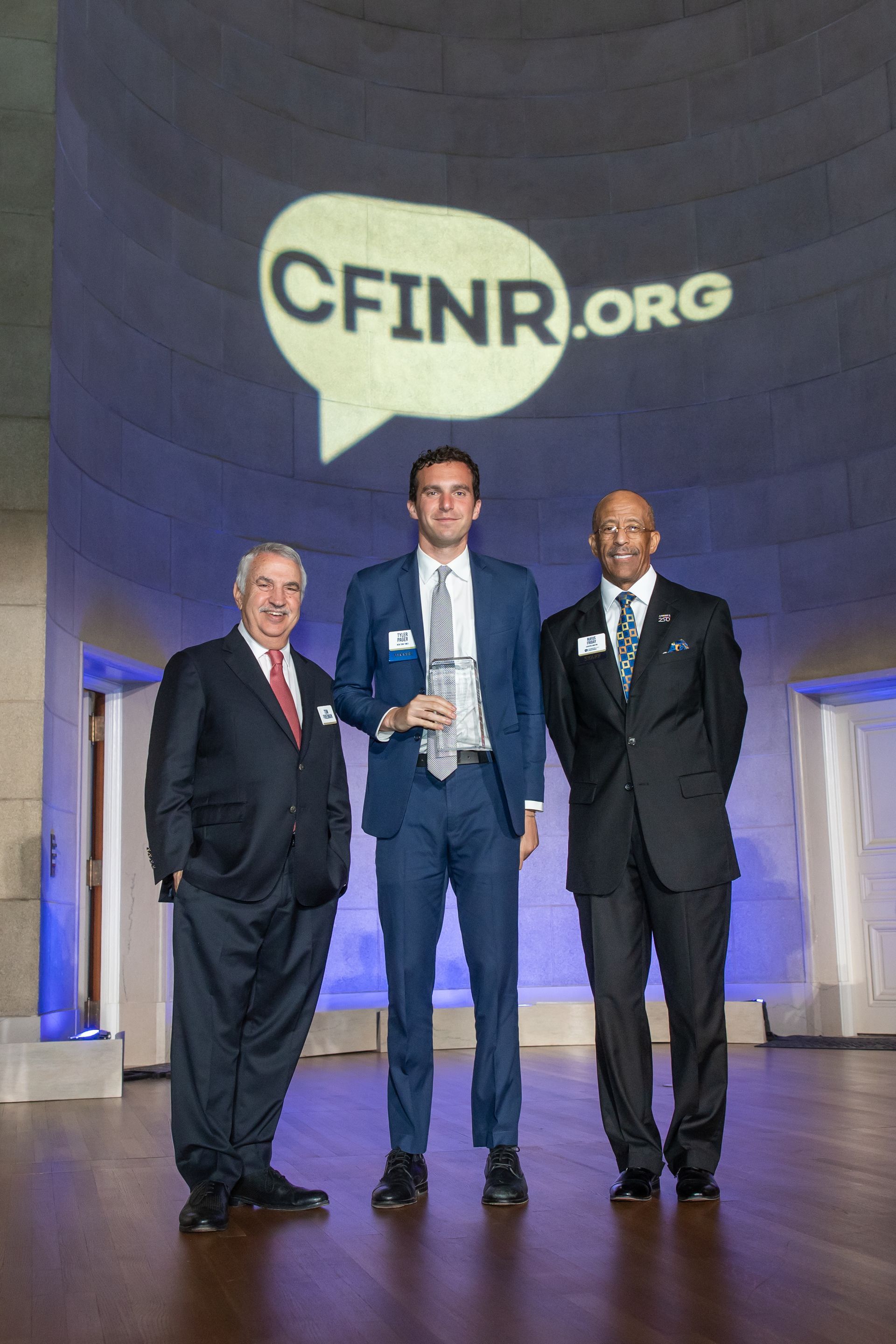 Three men in suits stand on a stage beneath a CFINR.org logo backdrop.