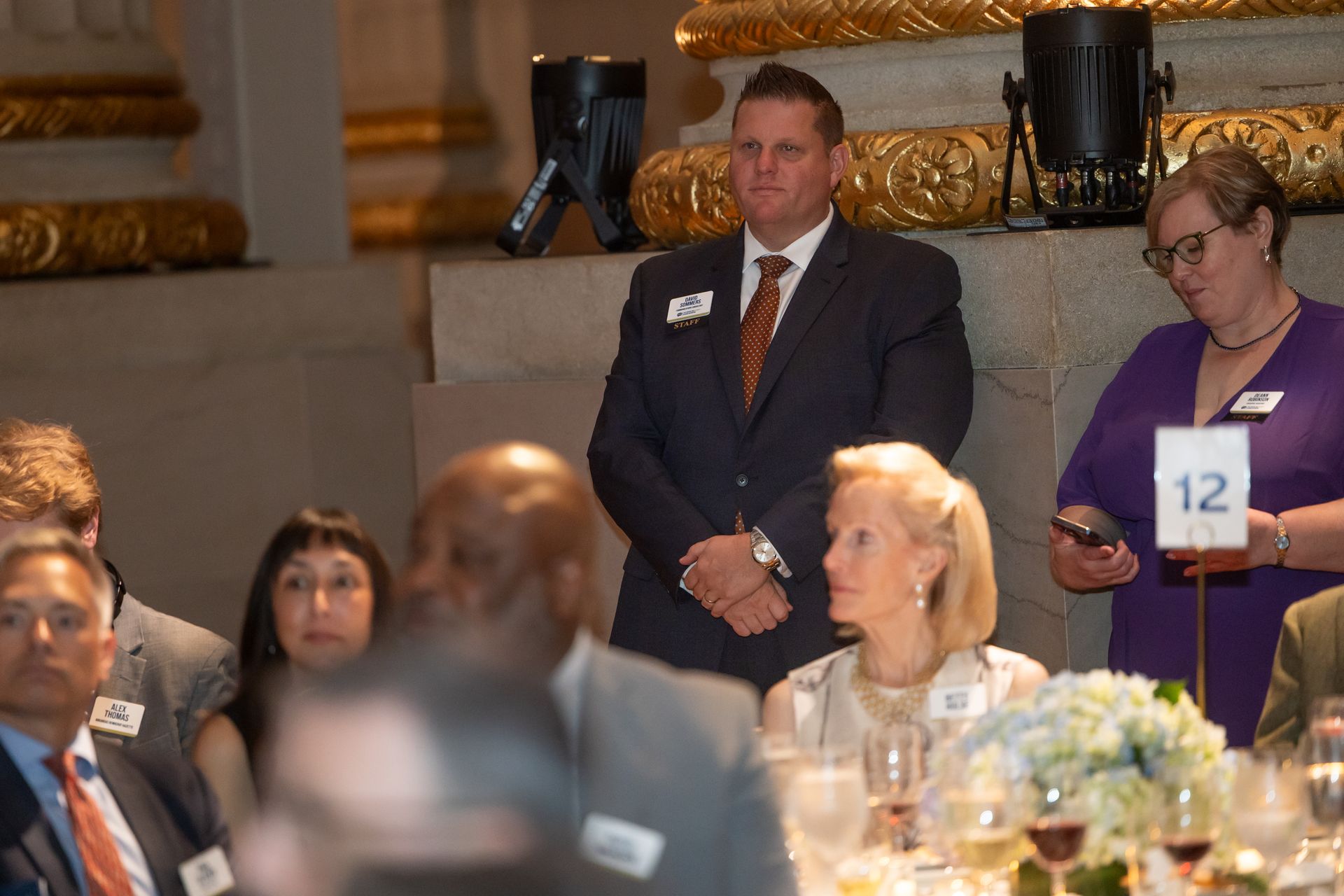 Man in a suit standing beside a woman in purple at a formal banquet table, holding a number 12 card