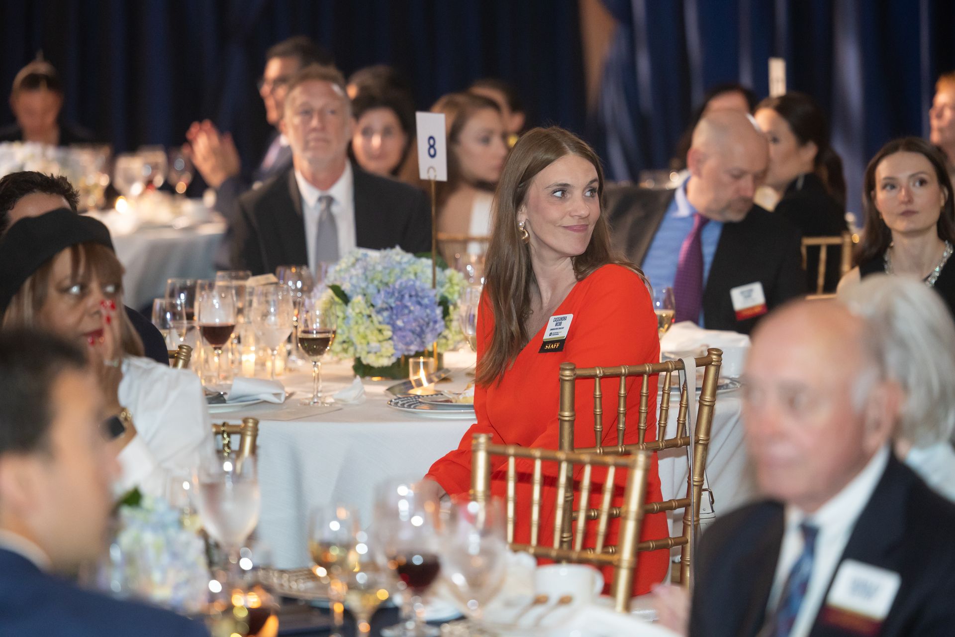 Guests seated at a formal banquet table, with a woman in a bright red dress in the center.