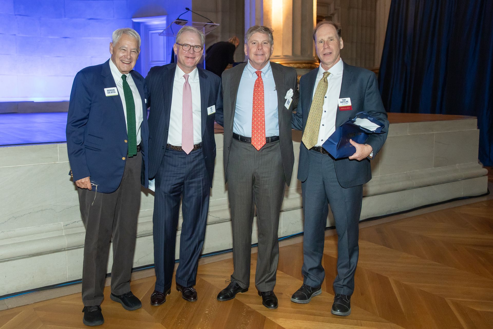Four men in suits pose together on a stage with blue curtains and wooden flooring.