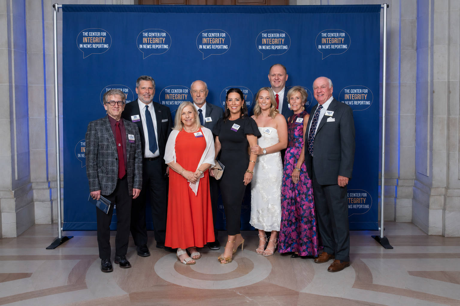 Eight people posing in formal attire before a blue step-and-repeat backdrop at an event.