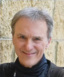 A man with gray hair is smiling in front of a stone wall.