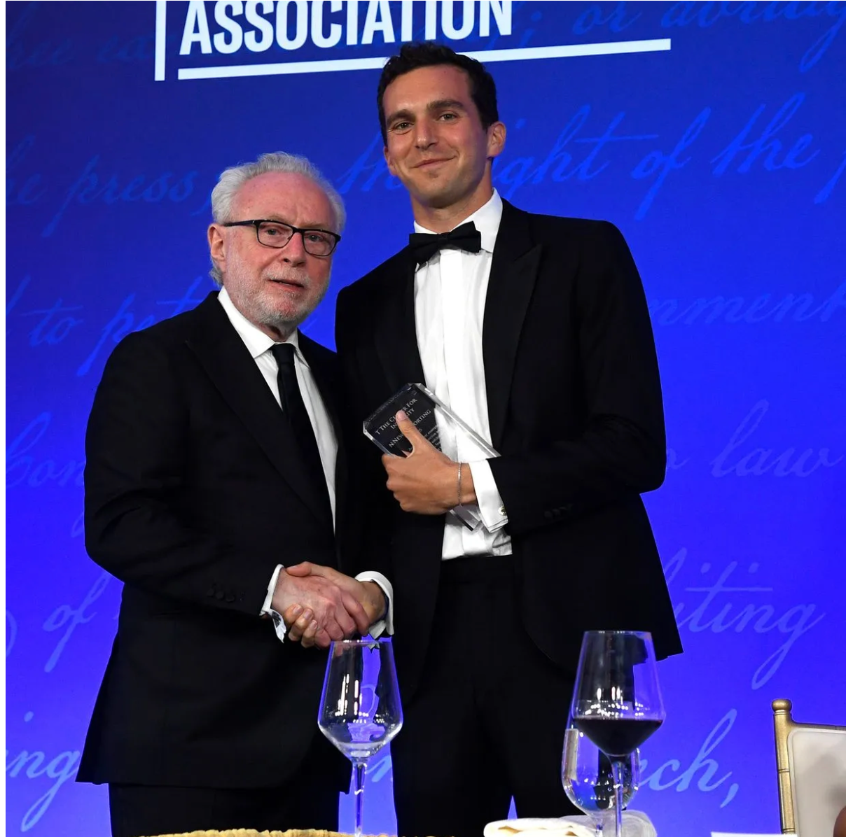 Three people on a stage at an awards event, holding trophies before a blue backdrop and U.S. flag.