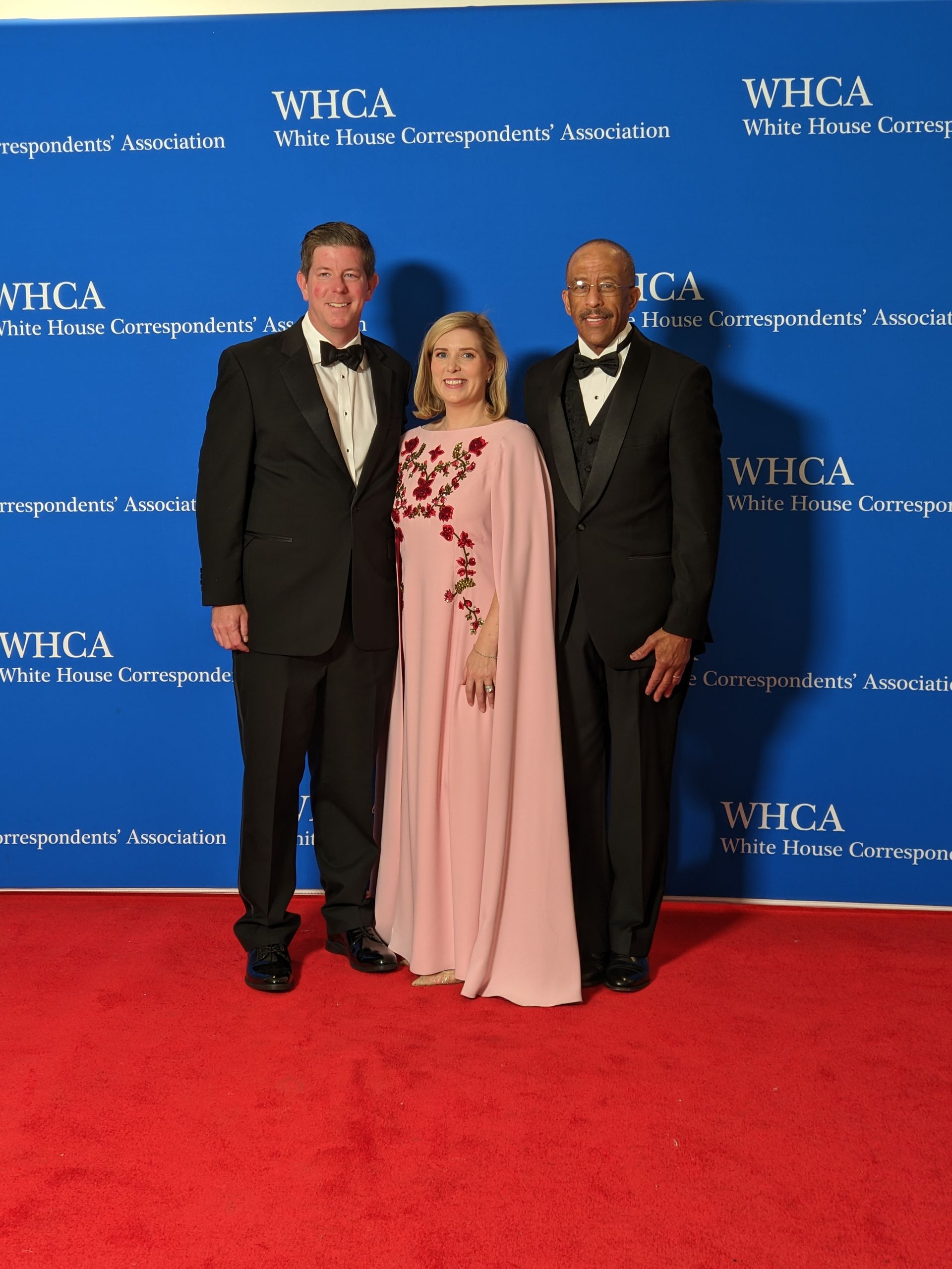 Three people standing on a red carpet in front of a wall that says whca