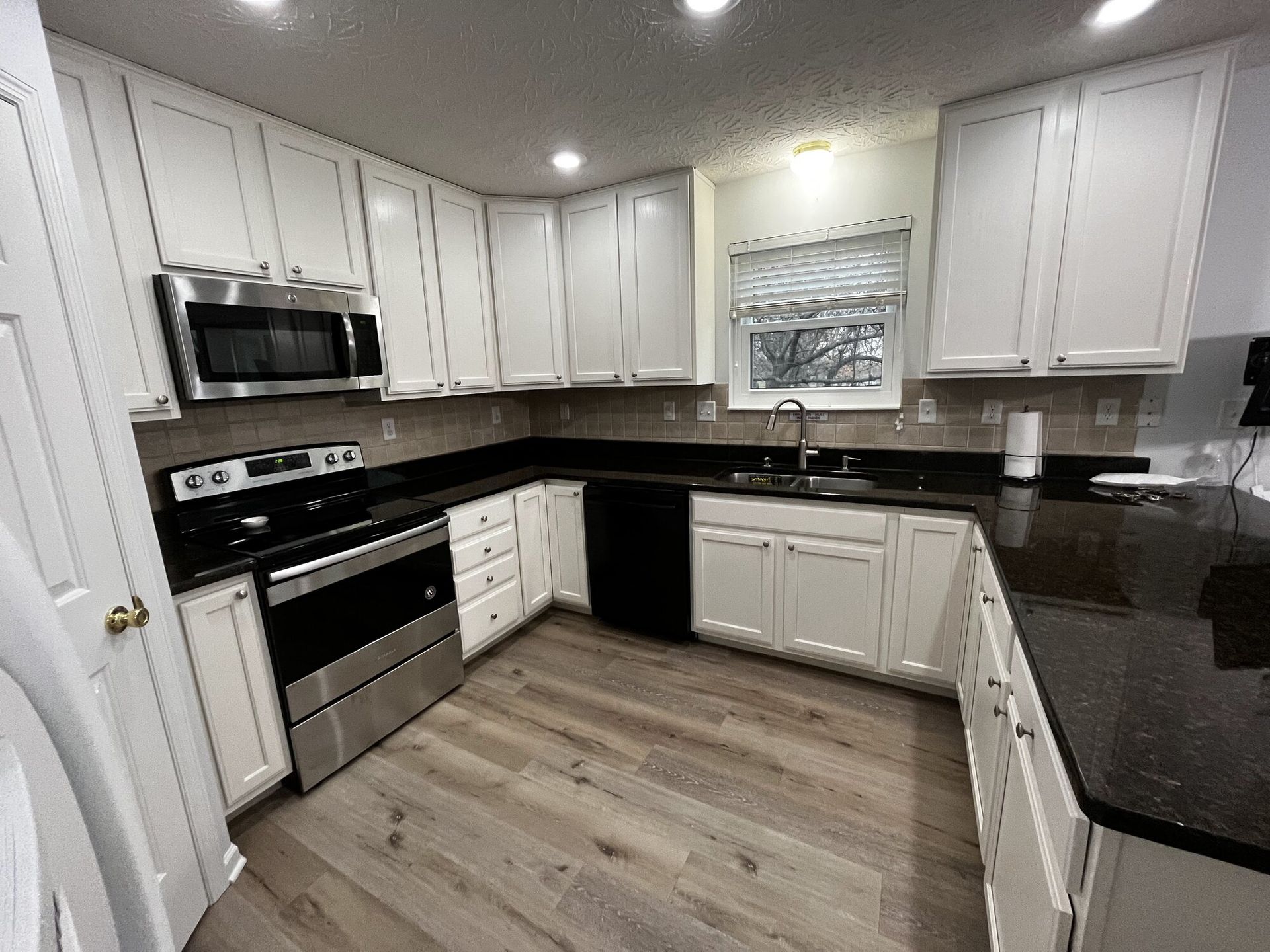 Bright modern kitchen with white cabinets, black countertops, stainless steel appliances, and wood-look floor.