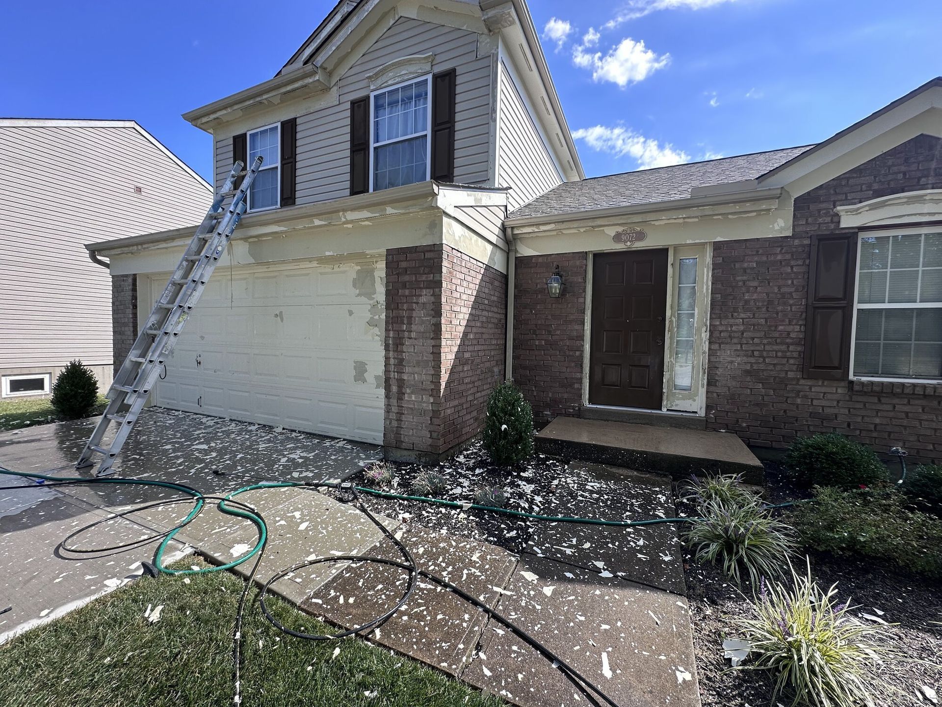 Damaged house exterior with a bent garage door, brick columns, and debris scattered across the driveway.