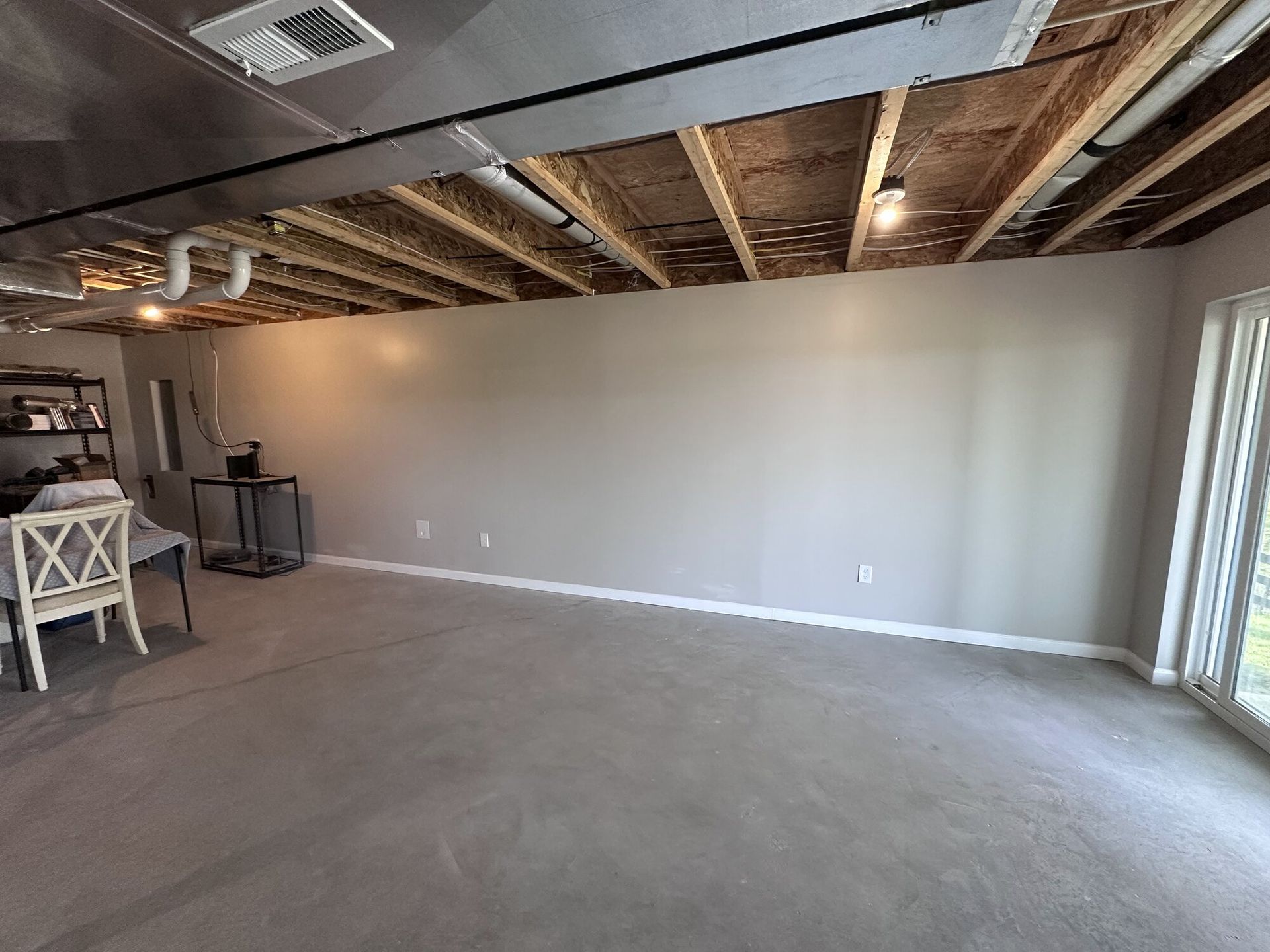 Empty unfinished basement room with exposed ceiling joists, gray carpet, and sliding glass doors.
