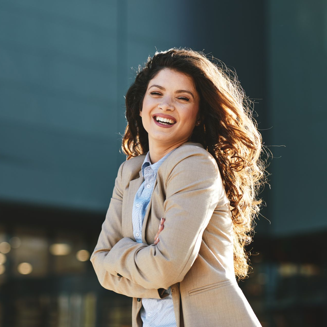 Smiling woman in a beige blazer standing outdoors in sunlight with a dark building backdrop
