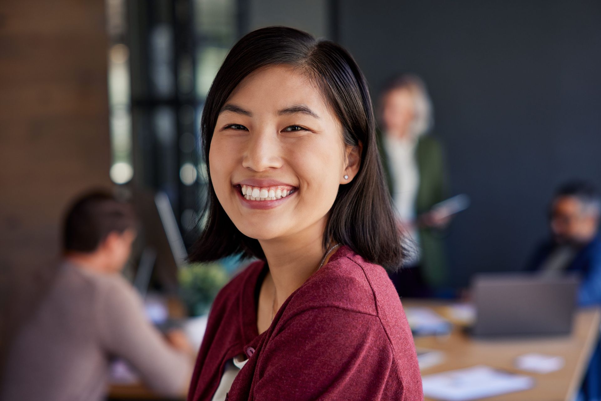 Smiling woman in a maroon sweater seated in a blurred office meeting room