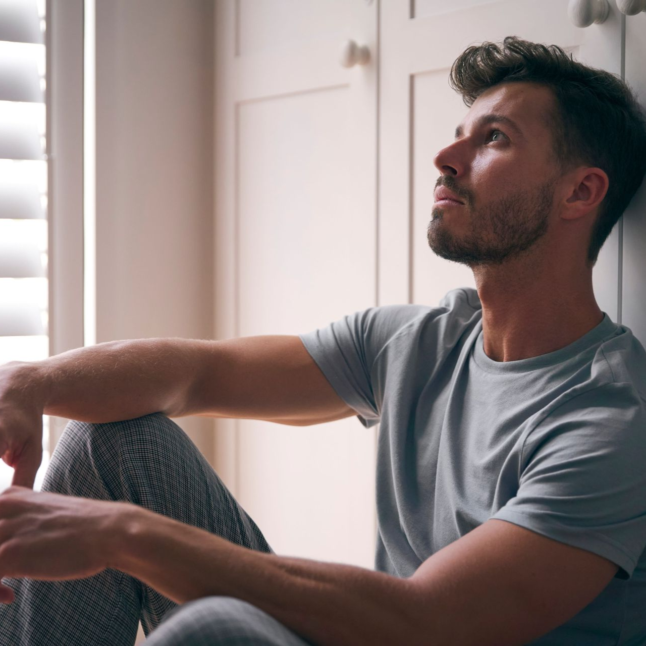 Man seated indoors by a window, looking upward in a gray T-shirt