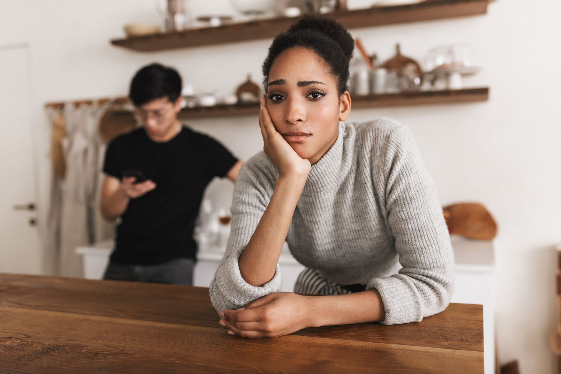 A woman leans on a wooden table, looking upset, while a man stands blurred in the background.