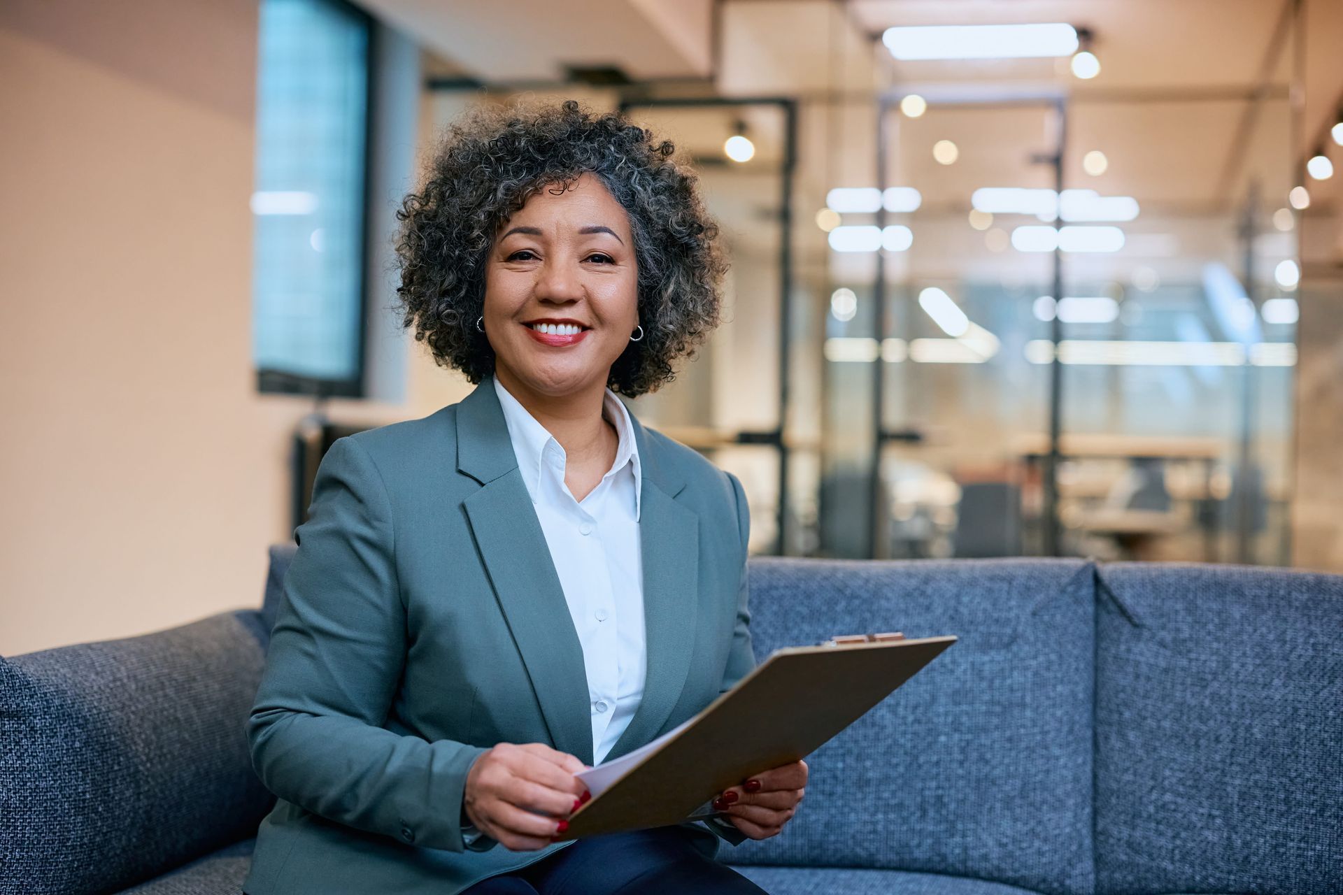 Person in a blue blazer holding a clipboard on a couch in a modern office lobby.