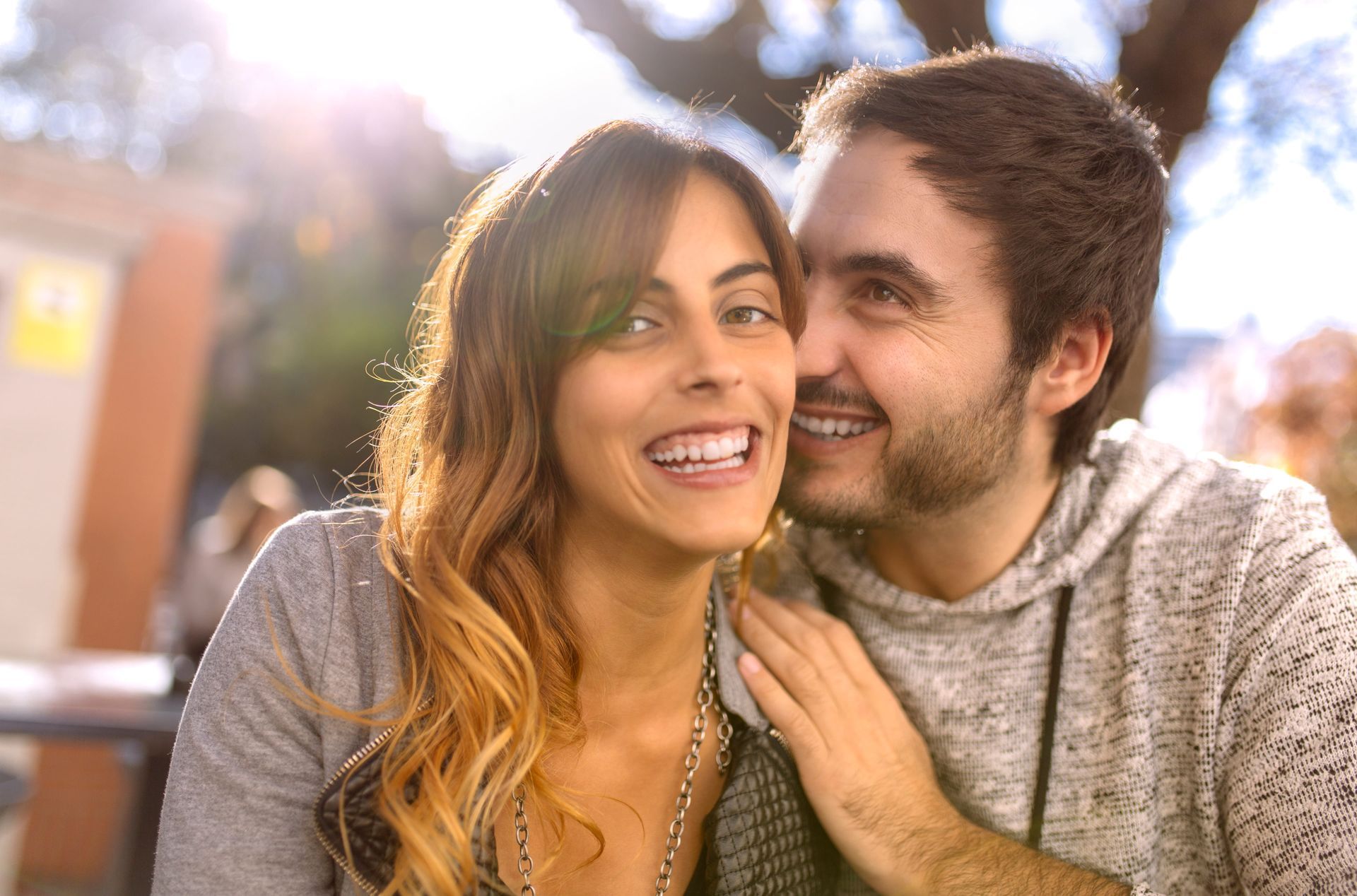 Smiling couple outdoors, leaning close in warm sunlight, with a blurred city background