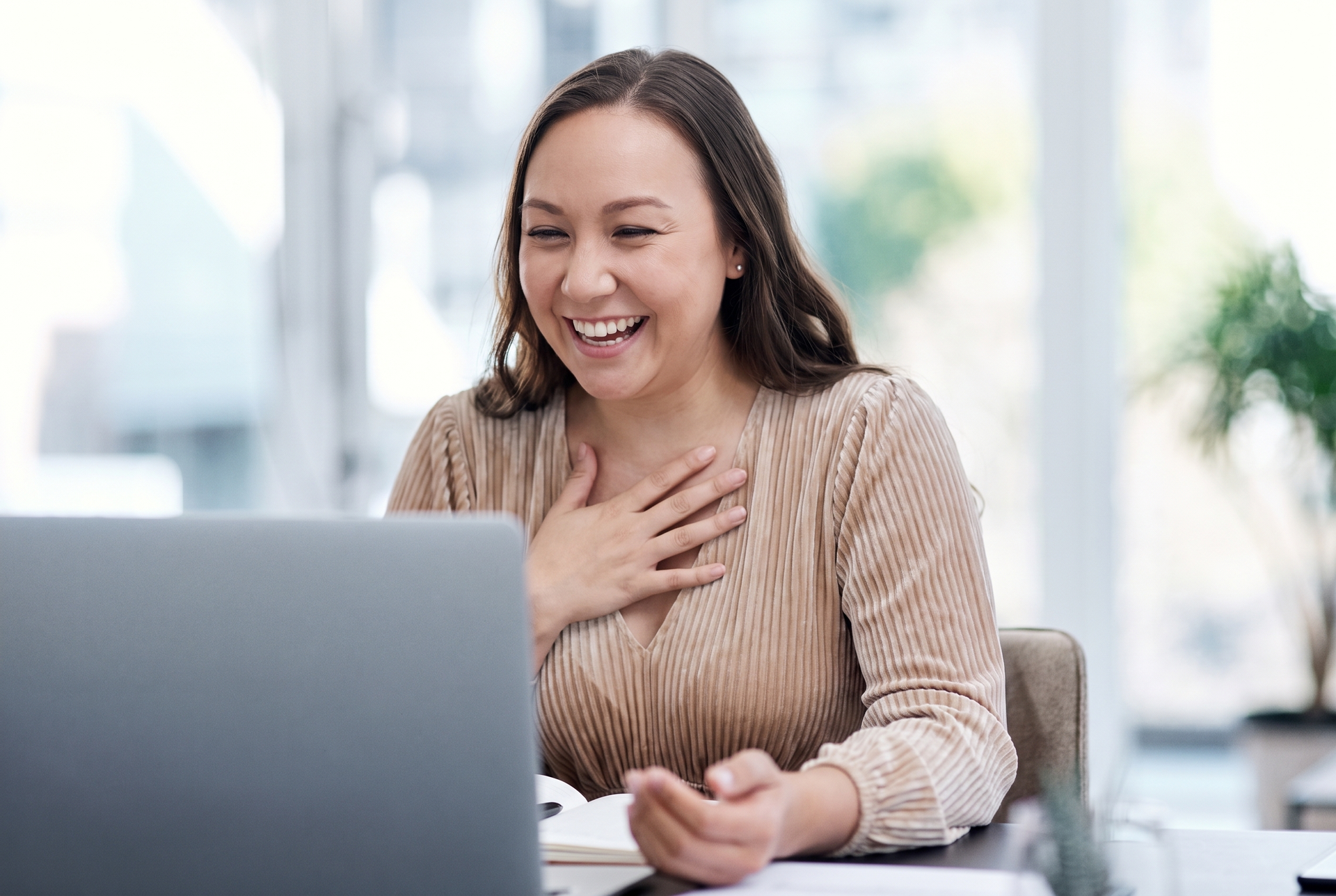 Smiling woman on a video call, hand on chest, sitting behind a laptop in a bright office