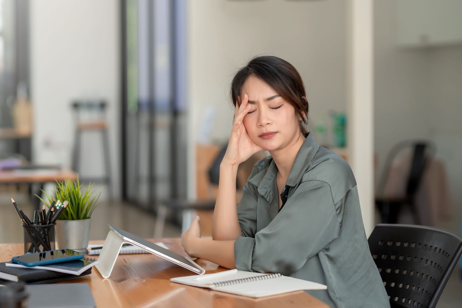 Woman at a desk, resting her head on her hand beside an open notebook and tablet in an office