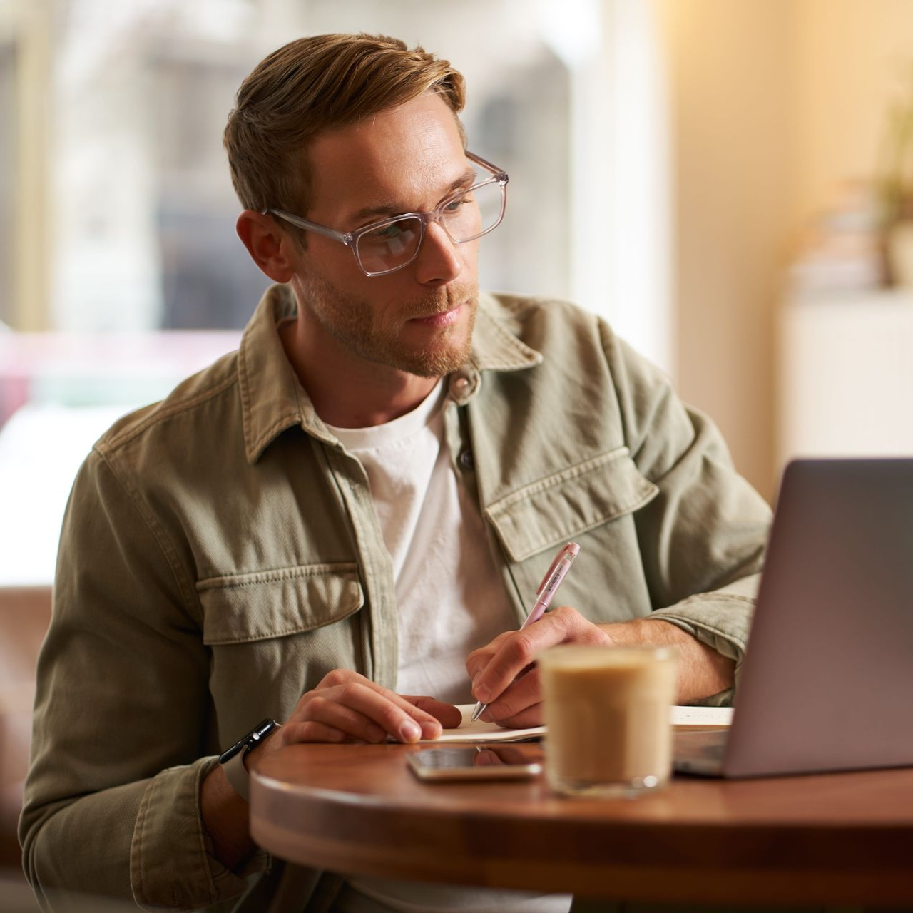 Man writing at a café table with a laptop and coffee