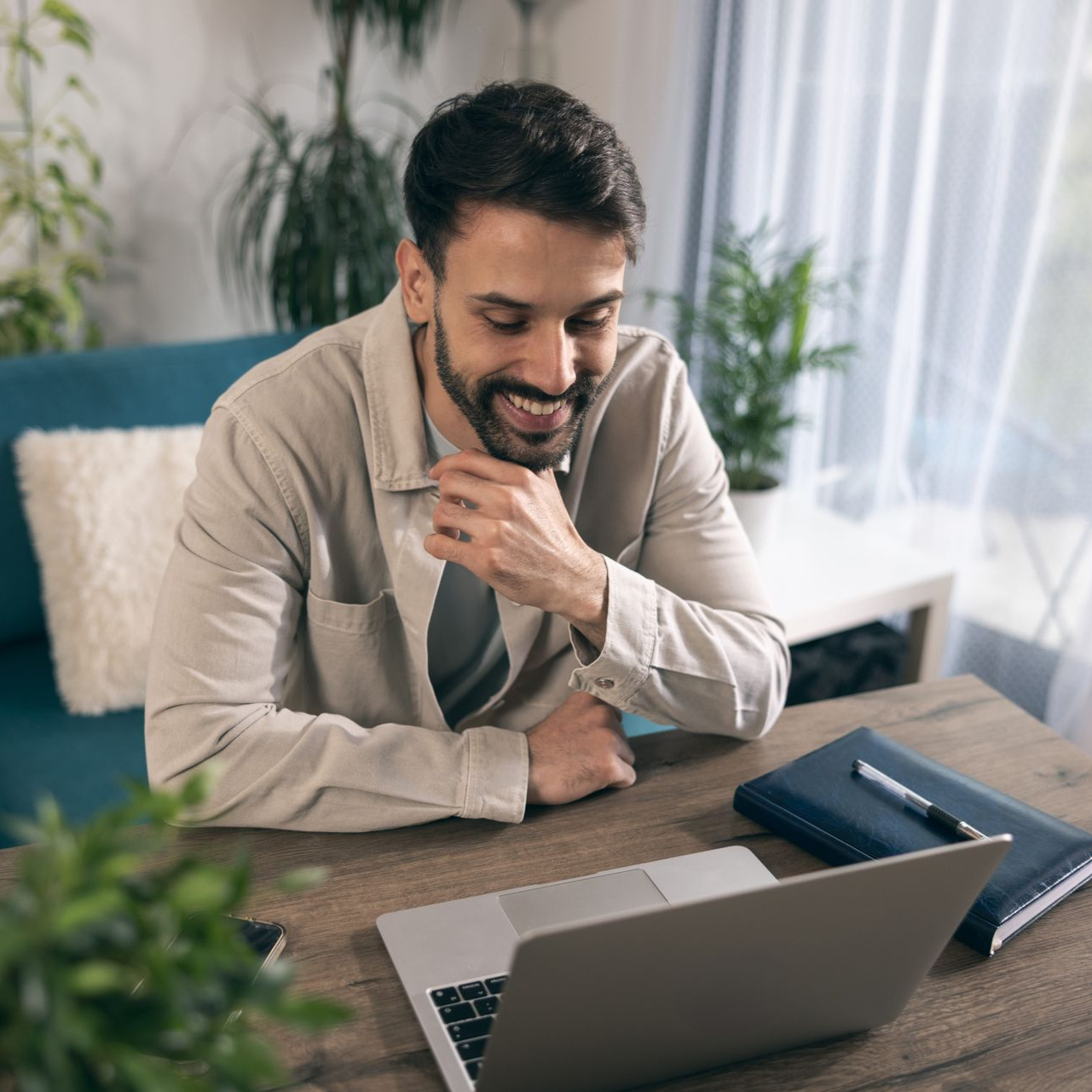 Man smiling at a laptop on a desk with a notebook and pen in a bright home office