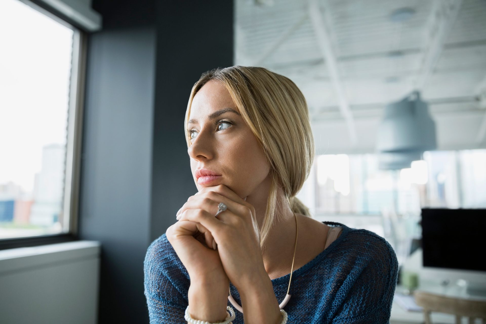 Woman in a blue sweater gazes out a window in a modern office, resting her chin on her hands.