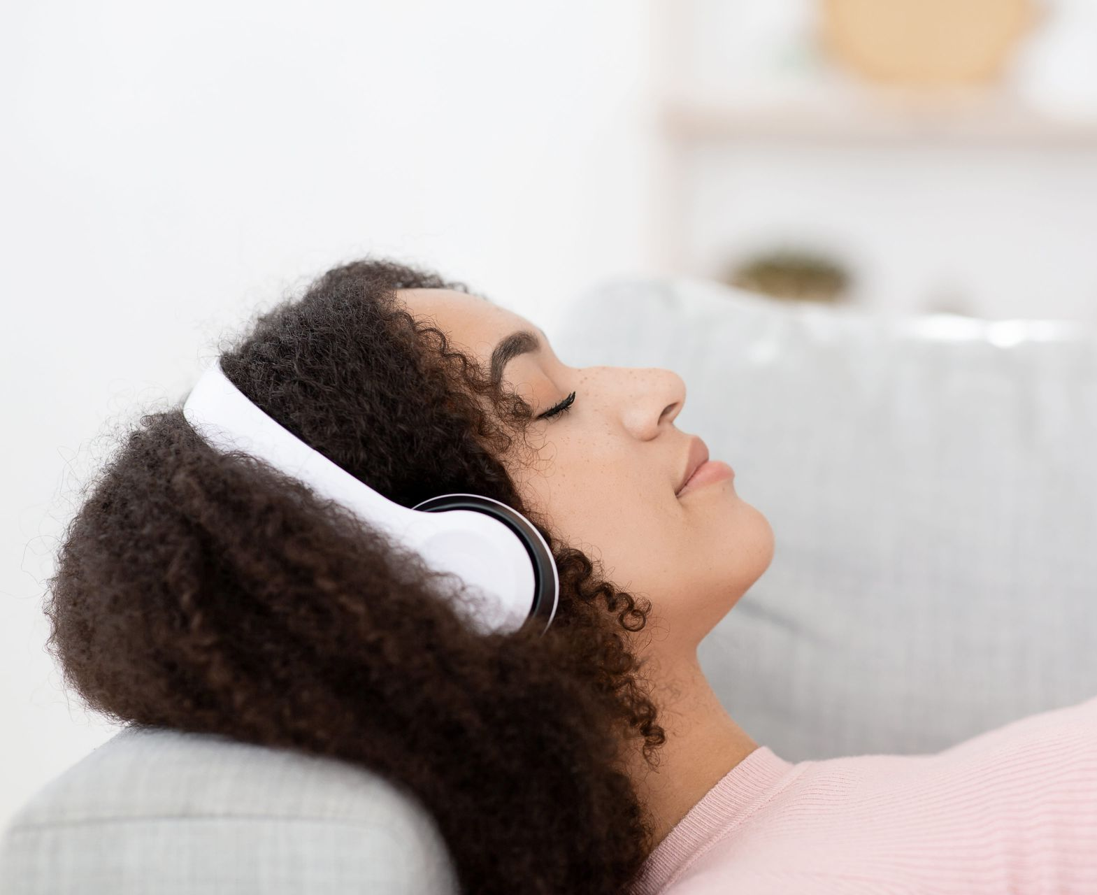 Person reclining on a couch wearing white headphones, with eyes closed in a bright, soft-lit room
