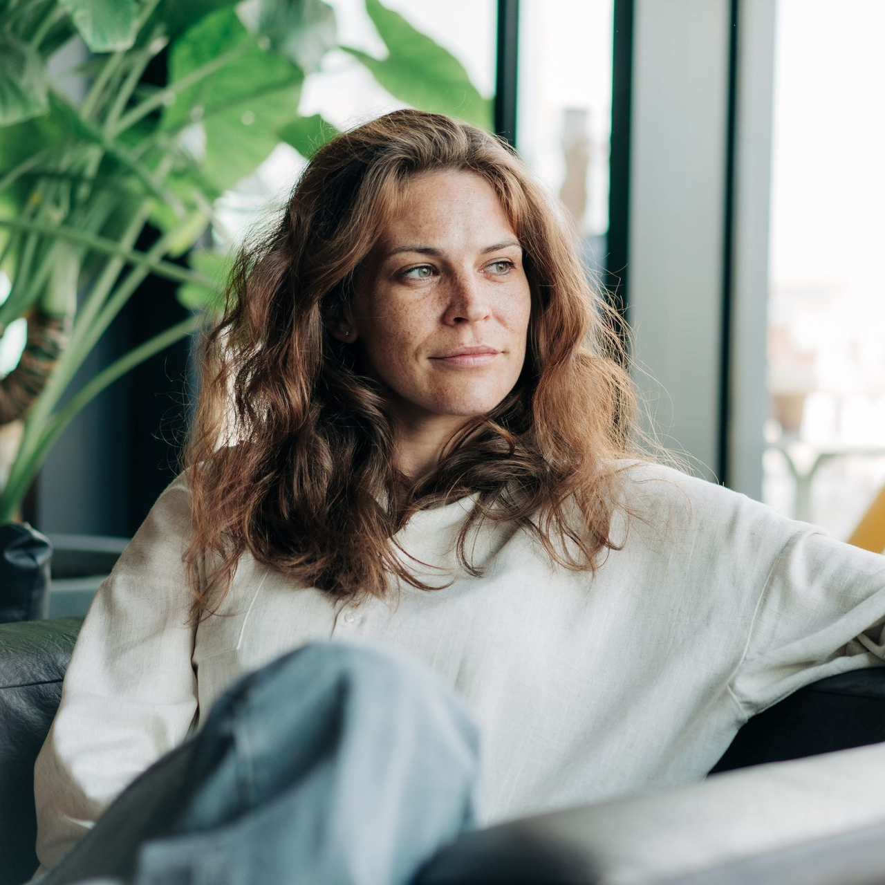 Woman sitting on a sofa by a window with green plants, looking to the side
