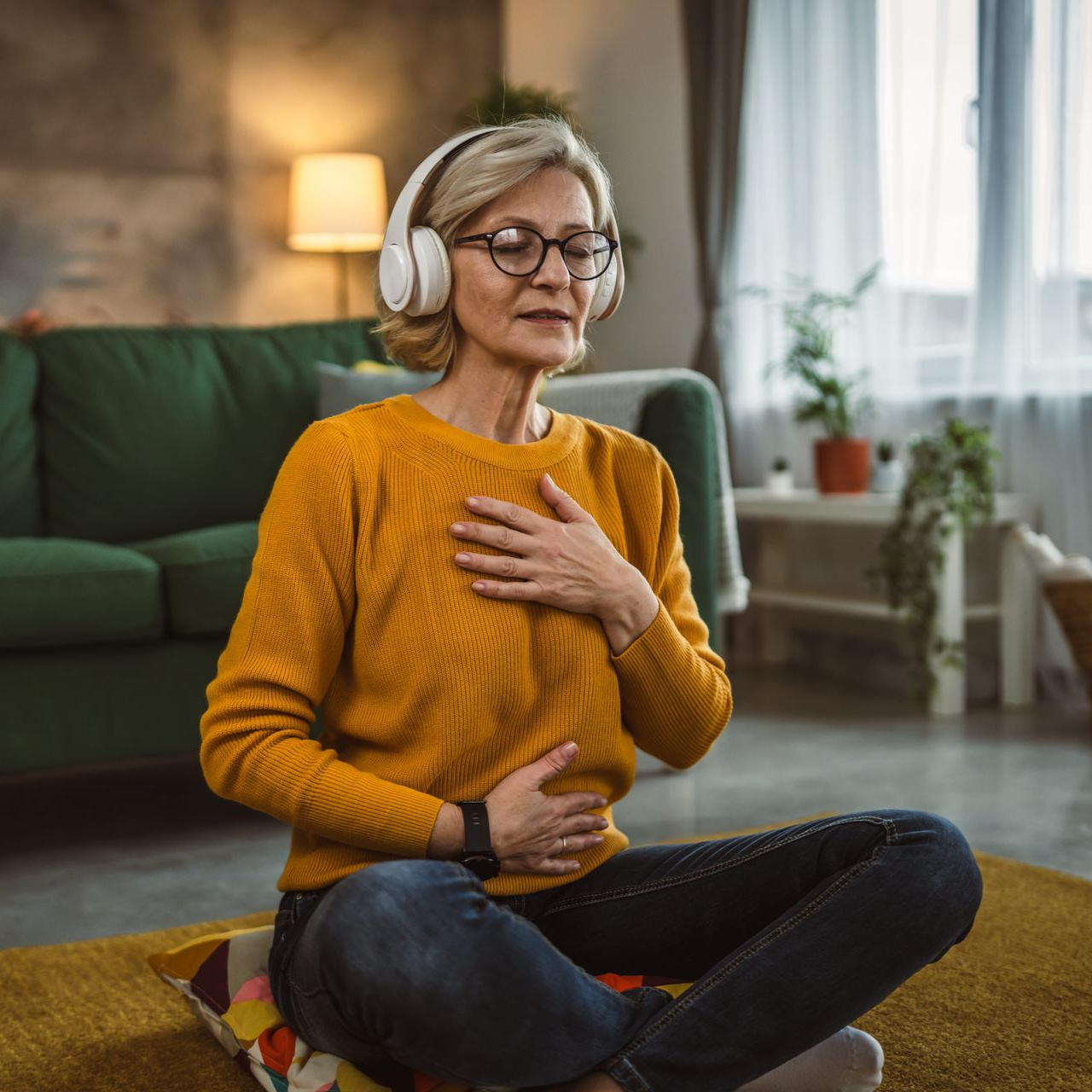 Person meditating indoors on a rug, wearing headphones and an orange sweater, with one hand on chest and one on stomach