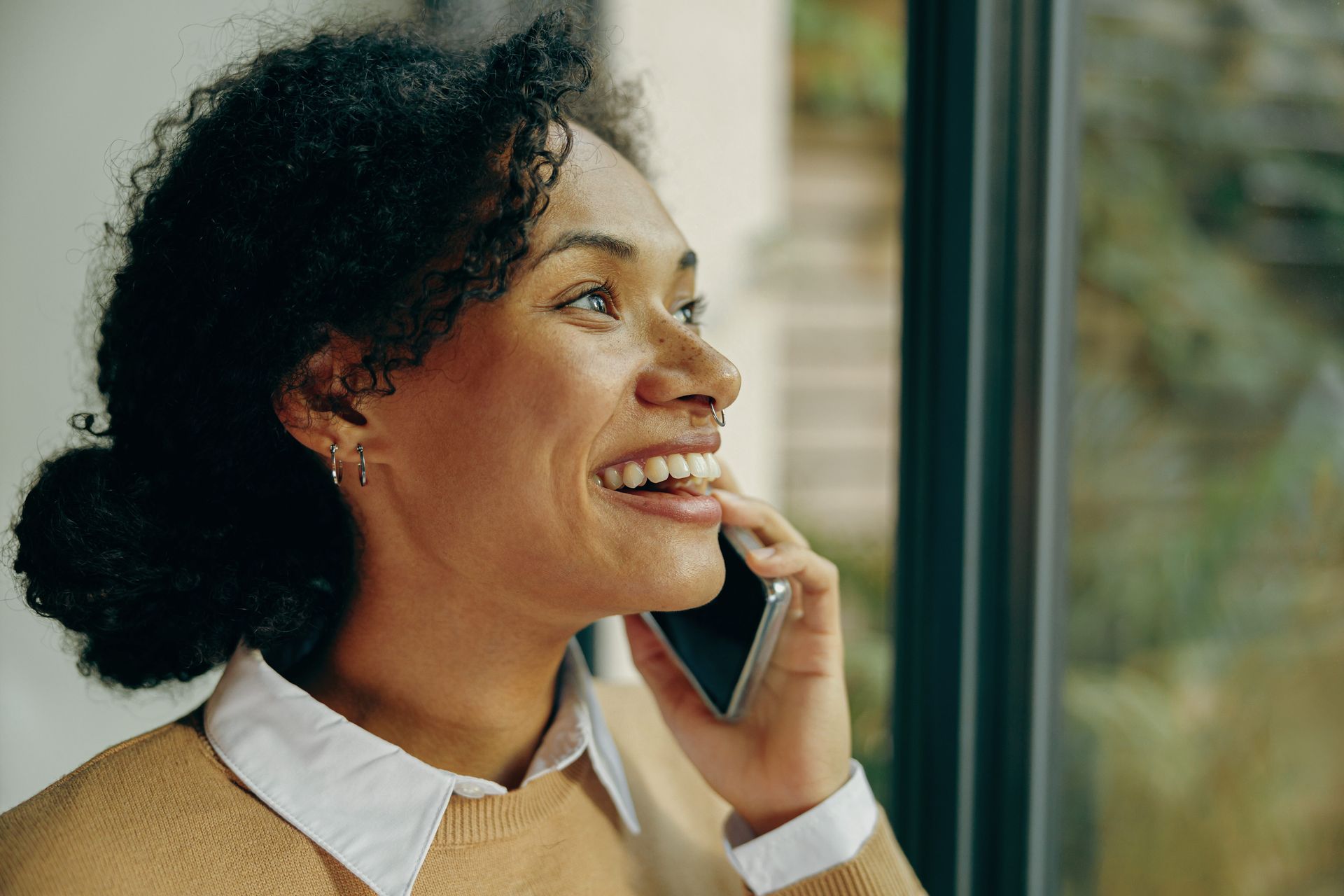 Smiling person talking on a smartphone by a window in a tan sweater and white-collared shirt