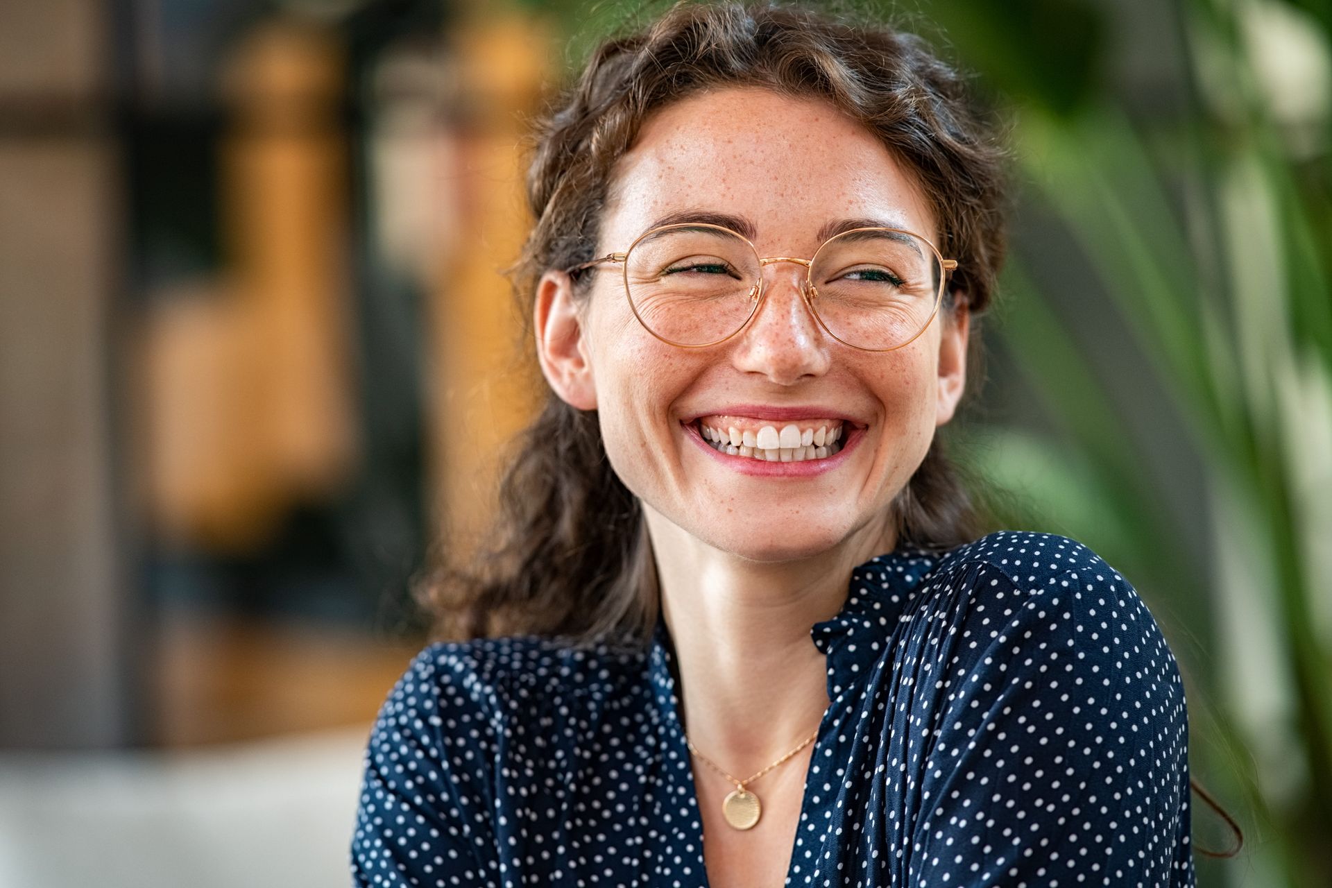 Smiling person with glasses in a navy polka-dot blouse, seated indoors with a blurred background