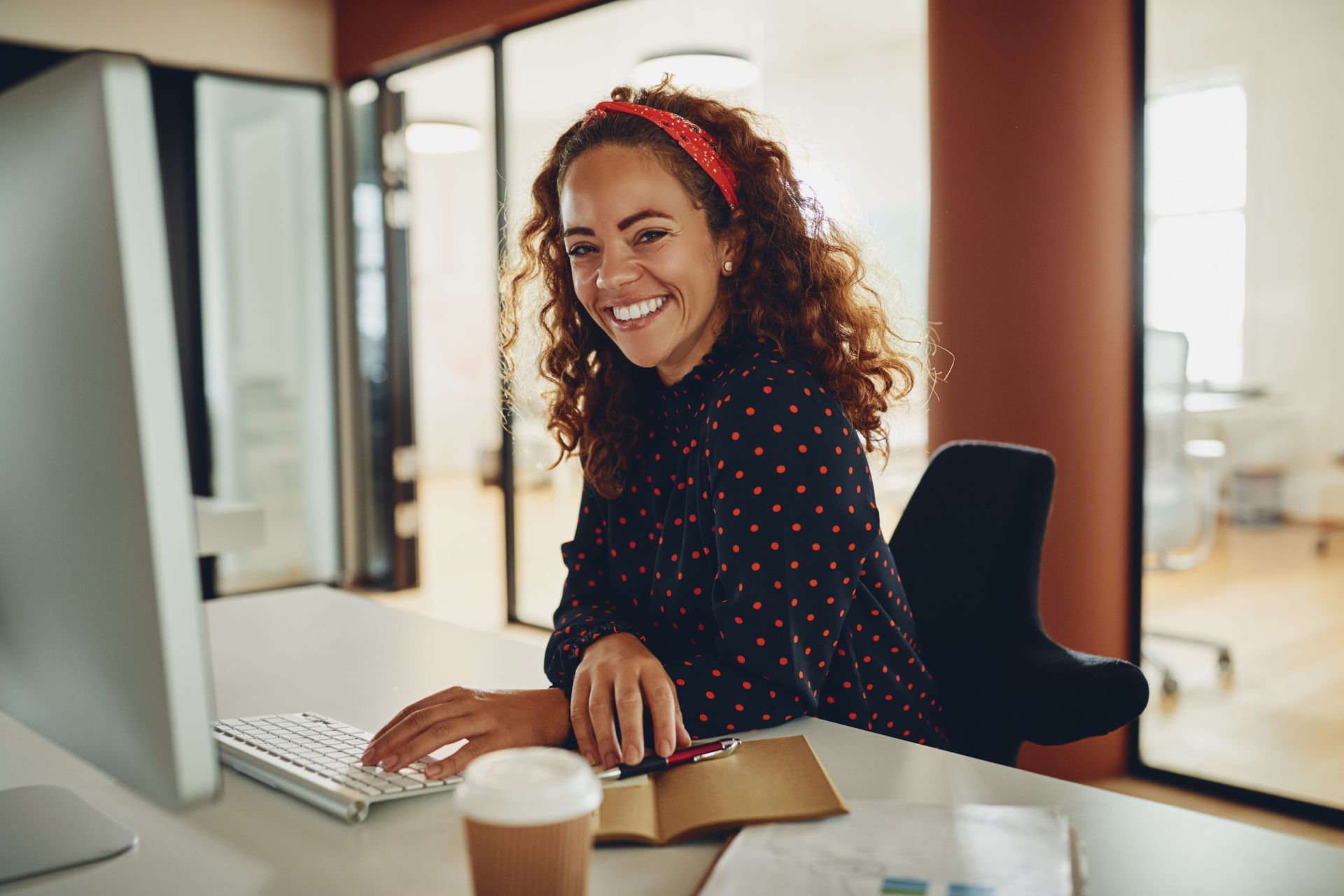Smiling person at a desk in a bright office, using a computer with a coffee cup nearby