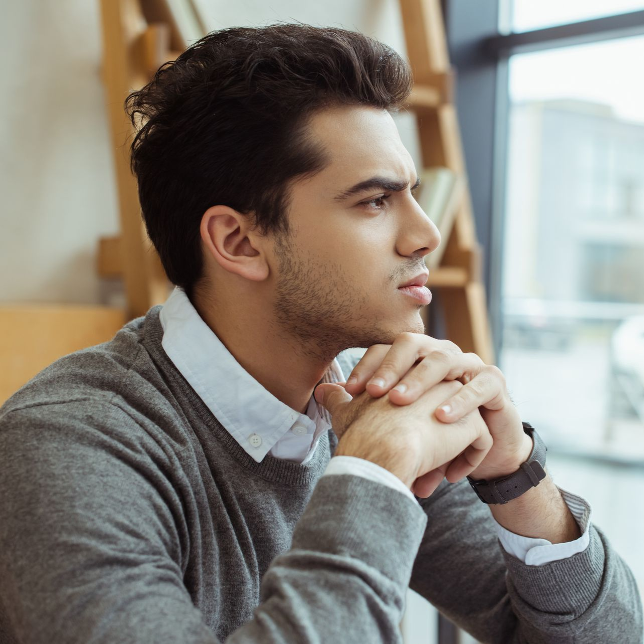 Man in gray sweater gazes out a window, hands clasped, in a bright room