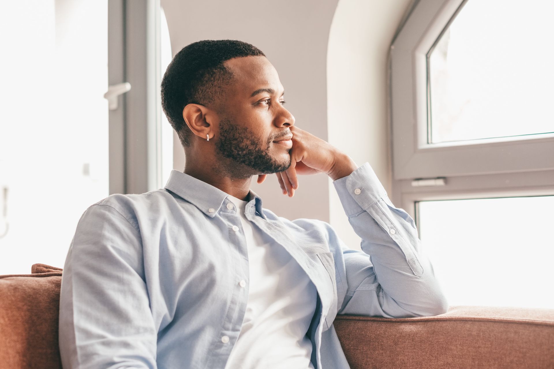 Man sitting on a couch by a window, looking thoughtfully outside.