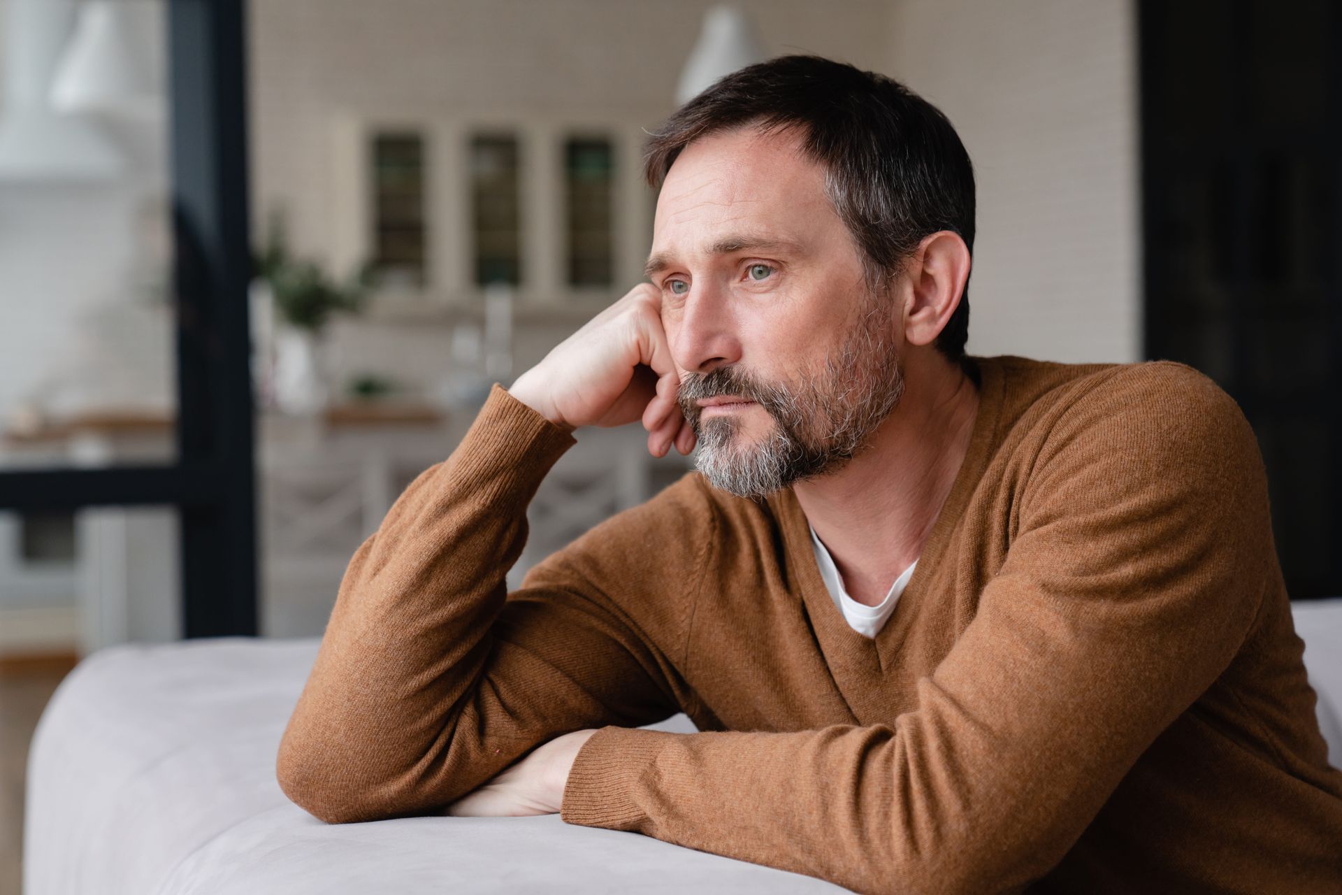 Thoughtful man in a brown sweater sitting on a couch, resting his head on his hand by a window.