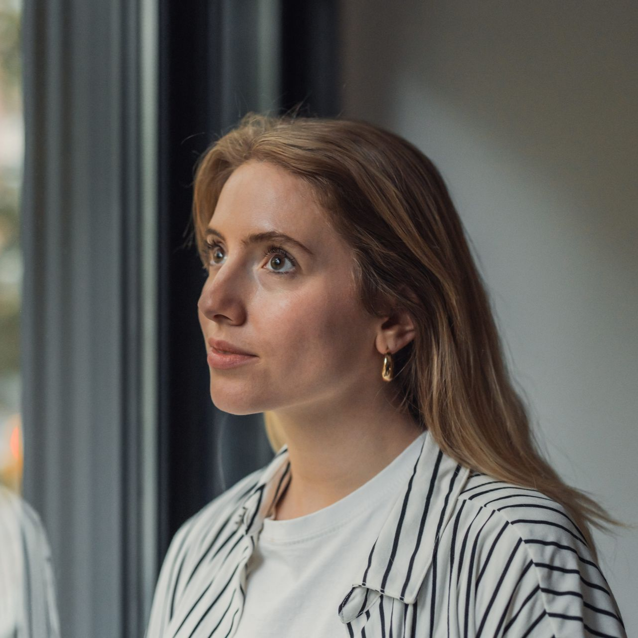 Woman in a striped shirt looking thoughtfully out a window indoors