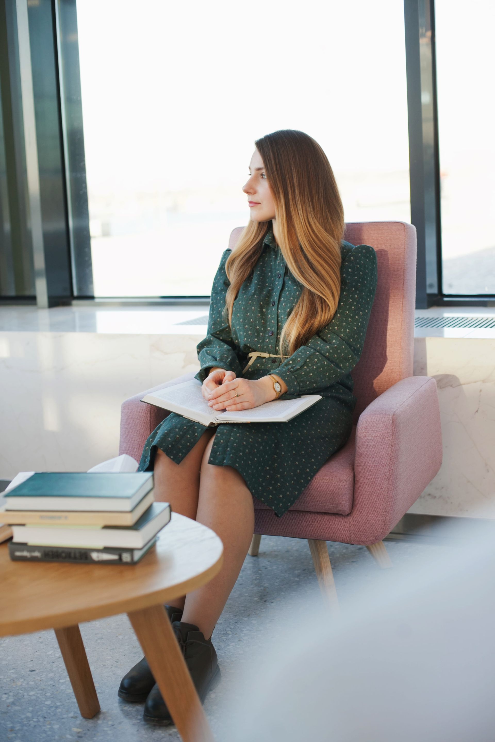 Woman seated in a pink armchair by a window, holding a notebook beside a coffee table with stacked books.
