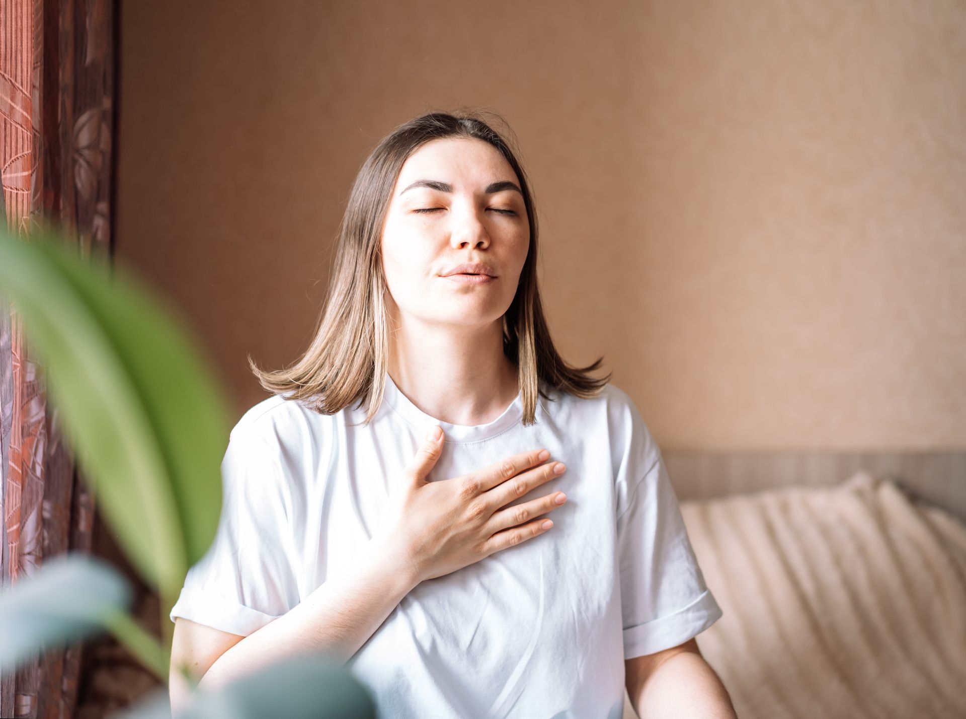 Woman in a white shirt sitting on a couch, eyes closed with hand on chest in a bright room