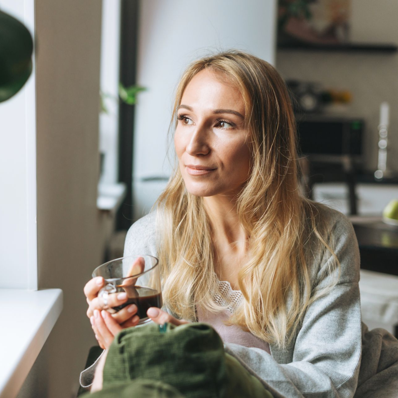 Woman sitting by a window holding a cup, looking out thoughtfully in a cozy room.