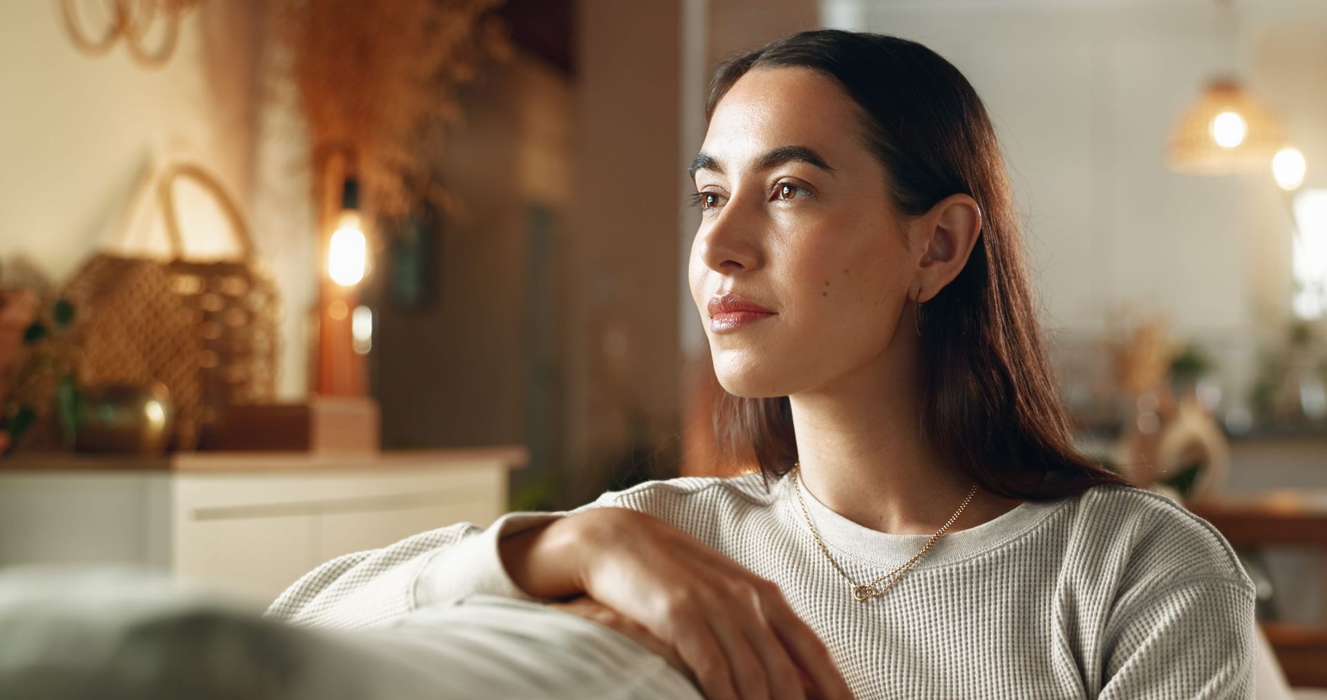 Person seated on a couch in a warmly lit living room, looking off to the side with a calm expression
