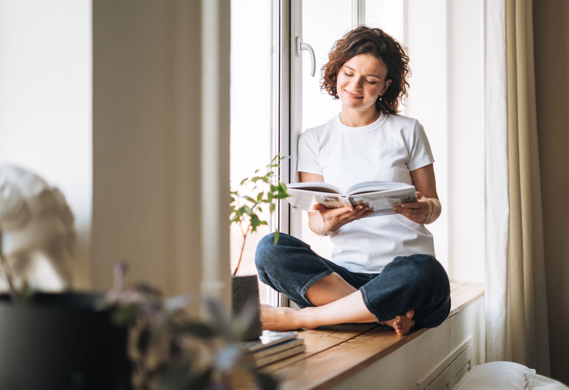 Woman sitting cross-legged on a windowsill, reading a book in a bright room.