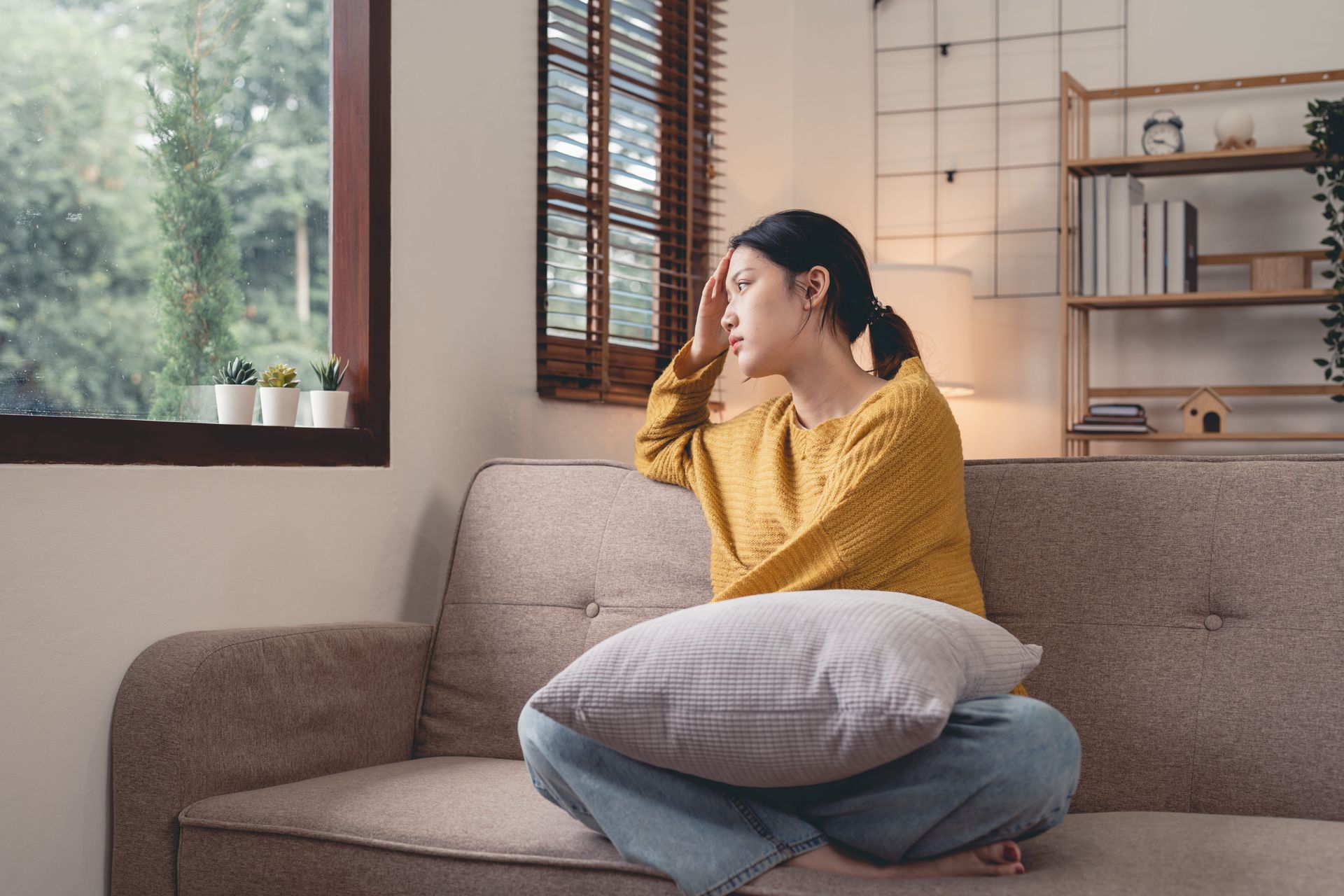 Woman sitting on a sofa with a pillow, looking out a window in a cozy living room