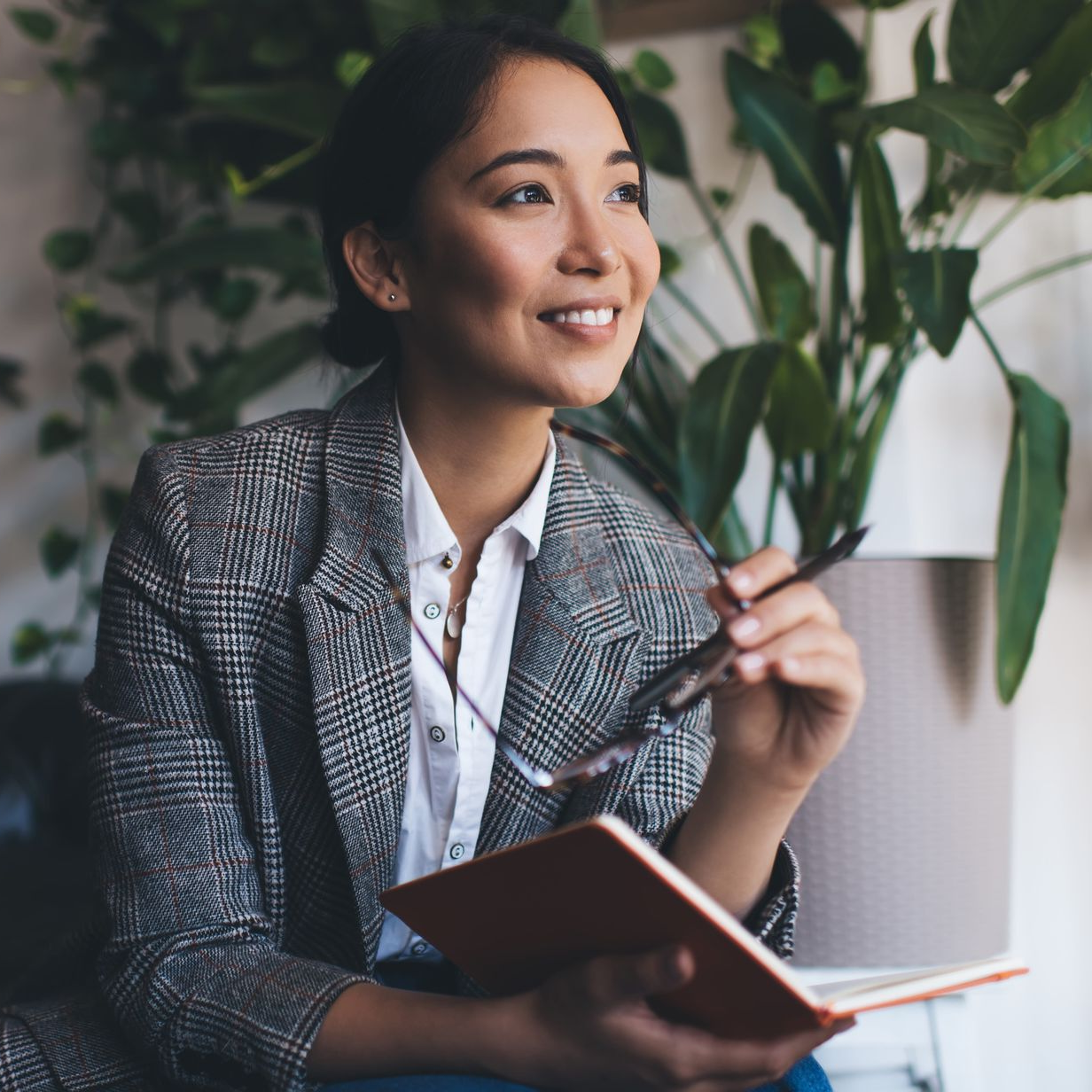 Smiling professional holding a notebook while seated indoors near a leafy plant