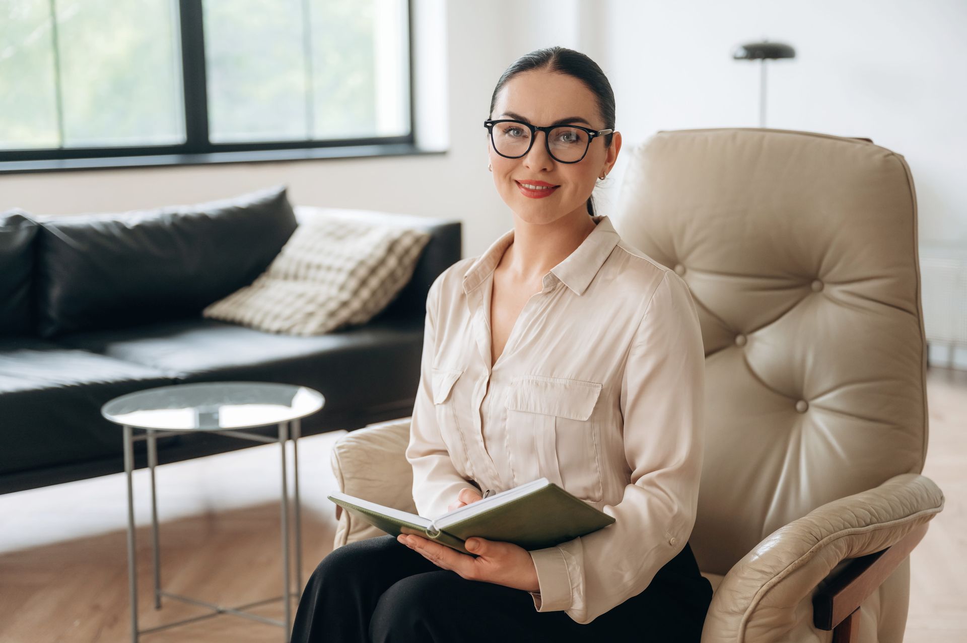 Woman with glasses sitting in a beige chair, holding a notebook in a bright living room.