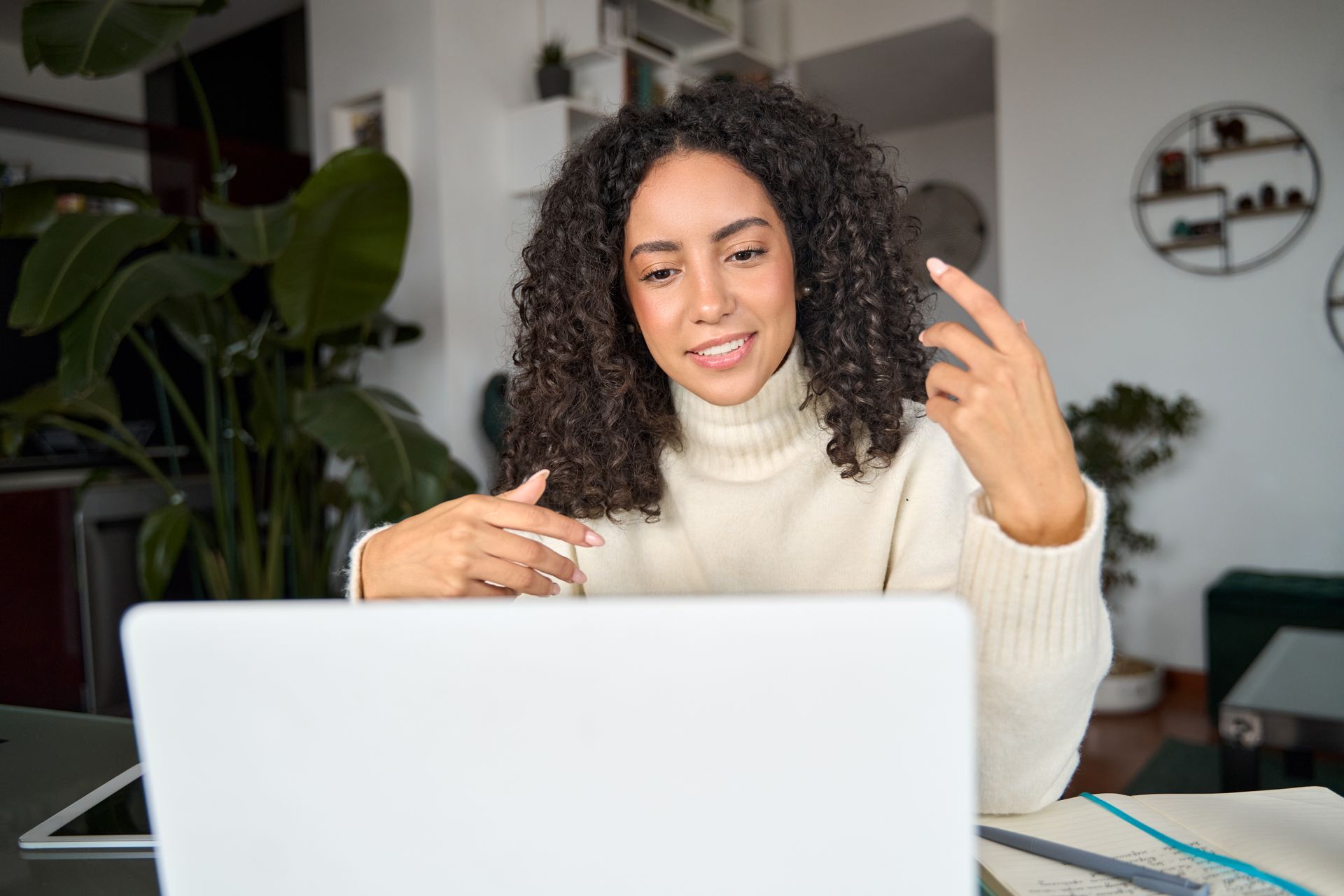 Woman on a video call at a laptop, smiling and gesturing in a bright home office.
