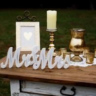 Rustic wedding table with “Mrs & Mrs” sign, framed heart art, candle, and gold decor