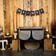 Rustic wedding sweetheart table with “MR & MRS” banner, wood backdrop, lanterns, and greenery accents