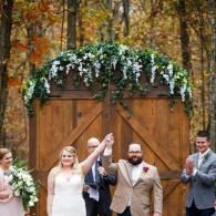 Wedding couple and guests celebrating under a floral arch in a wooded outdoor setting