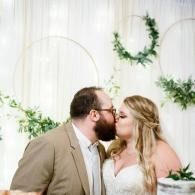 Two people kissing at a wedding, with greenery and white decor in the background