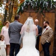 Wedding party gathered outdoors near wooden doors and greenery, with the bride in a white veil center.