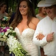 Bride in white dress walking arm-in-arm with a man in a white cowboy hat, holding a bouquet outdoors.