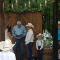 Outdoor gathering with people in cowboy hats standing near a wooden backdrop and white floral arrangement.