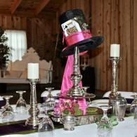 Decorated table centerpiece with pink ribbon, candles, and a tilted black top hat in a rustic room