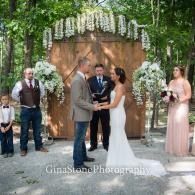 Wedding ceremony under a flower-covered arch with couple exchanging vows, officiant, and guests outdoors